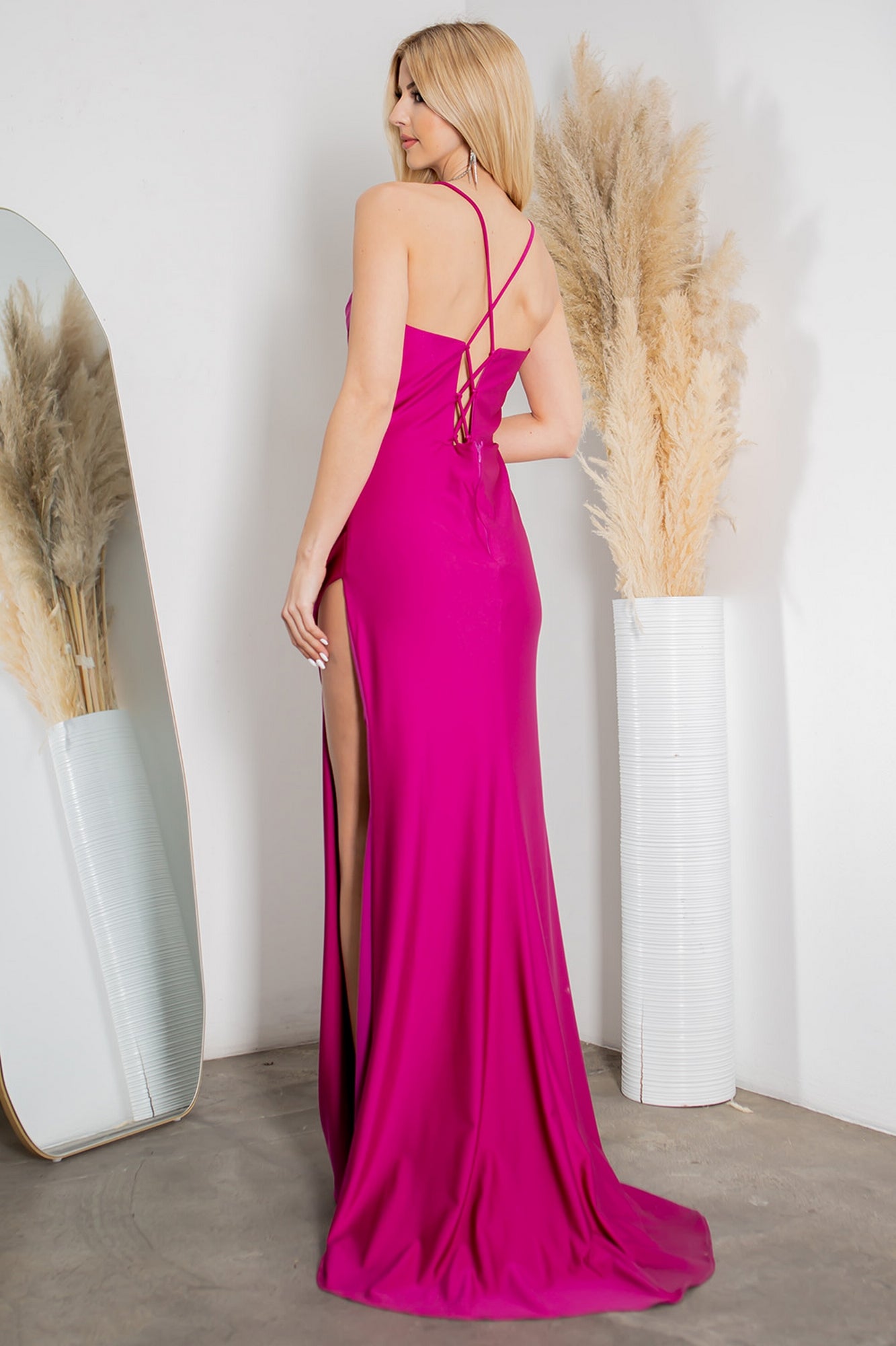 A woman models the Cowl-Neck Long Formal Corset Prom Dress 8035J—a bright pink, floor-length gown with a side slit and crisscross straps—posing with her back to the camera beside a tall vase of pampas grass.